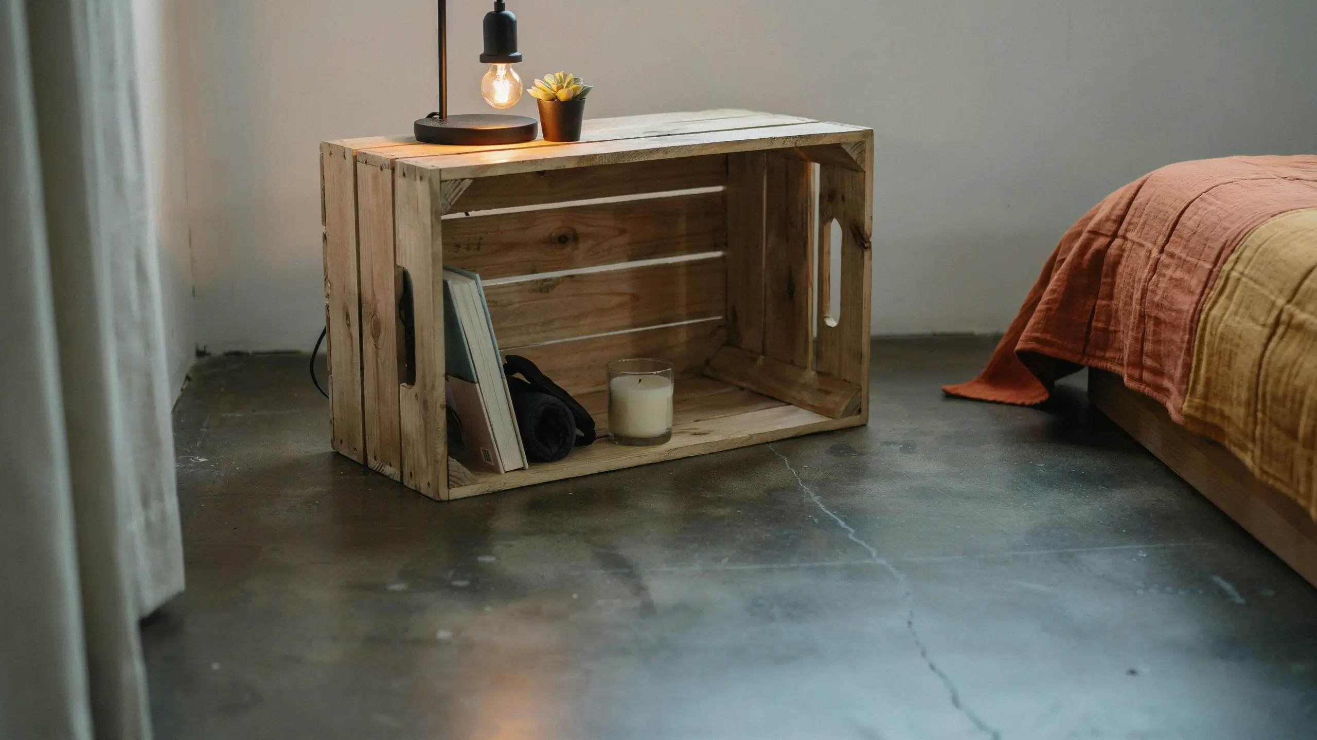 Rustic repurposed wooden crate used as a bedside nightstand, holding a vintage black lamp, small succulent, book, and white glass candle in a minimalist bedroom.