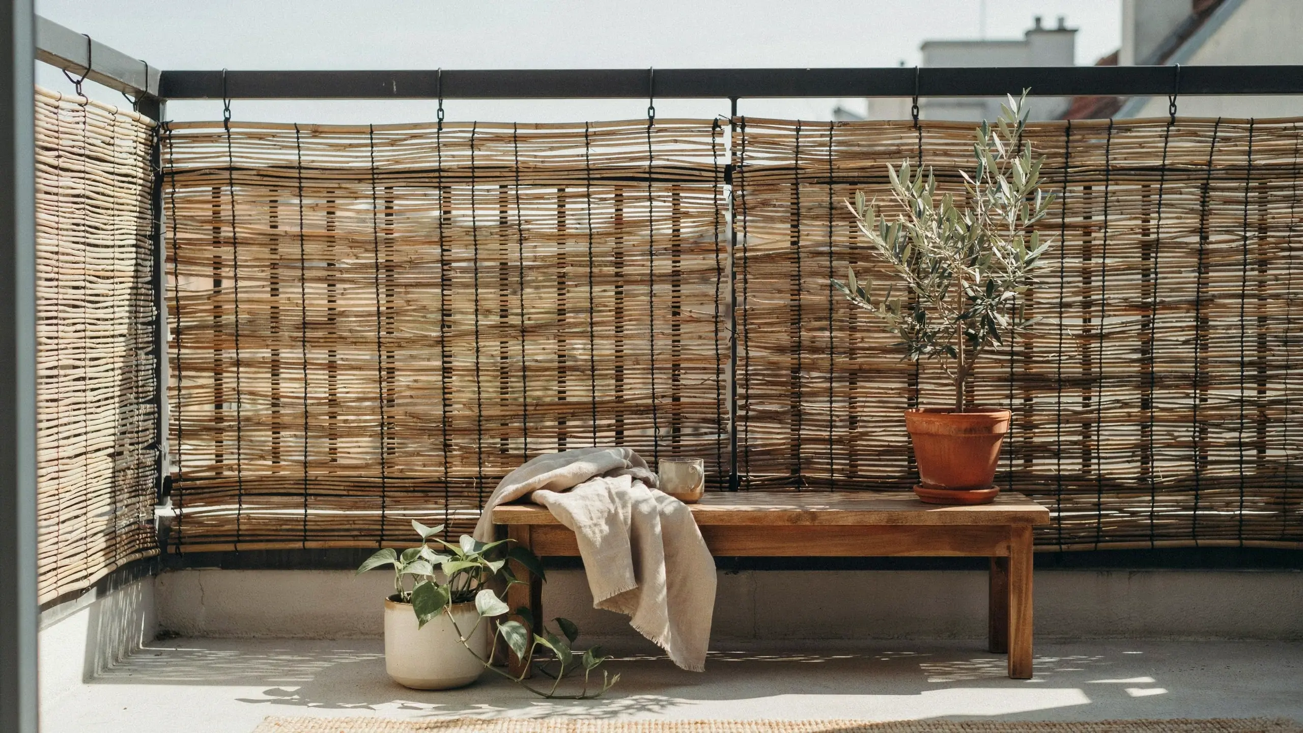 Woven bamboo and reed privacy screen attached to a black metal balcony railing, next to a wooden bench with a cosy throw blanket and a potted olive tree.