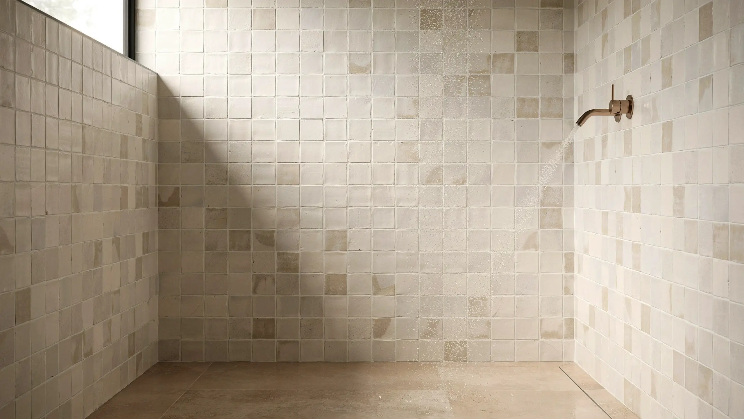 Handmade square cream zellige tiles in a minimalist walk-in shower with a wall-mounted brass showerhead and flowing water.